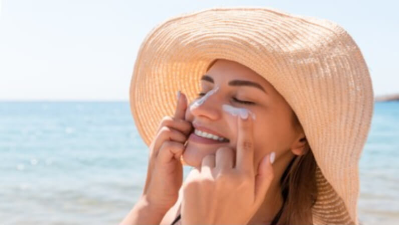 Woman on a beach wearing straw hat applying sunscreen on her face